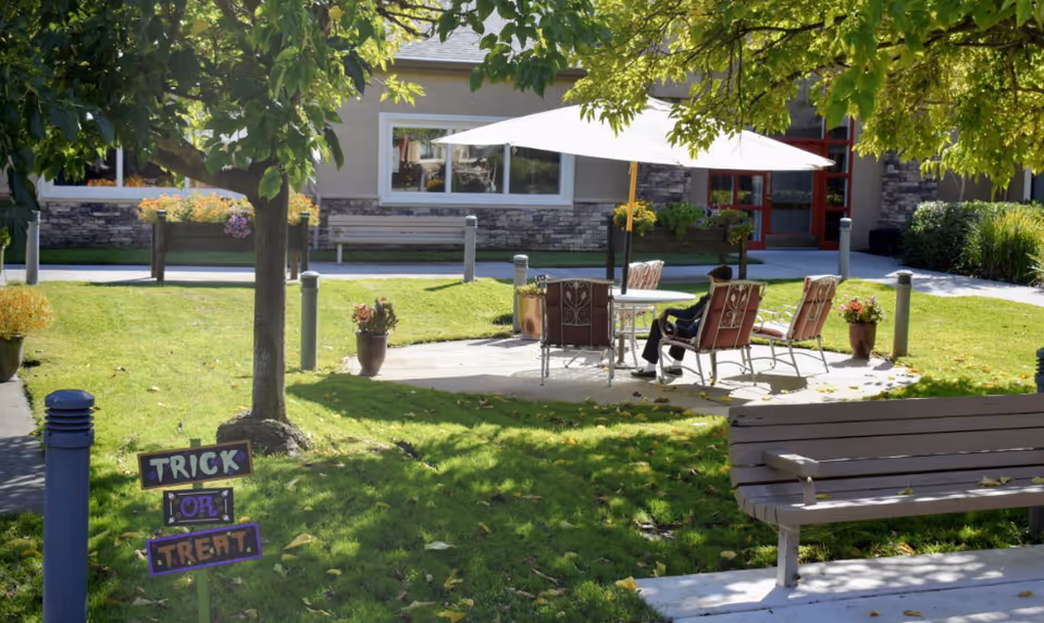 Sunny courtyard at Fair Oaks Estates with a patio table and umbrella, chairs, bench, trees, and a small "Trick or Treat" sign.