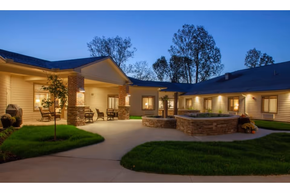 Dusk view of a lit courtyard with a covered patio, seating, stone planters/fountain, and landscaped walkways at a senior living facility.