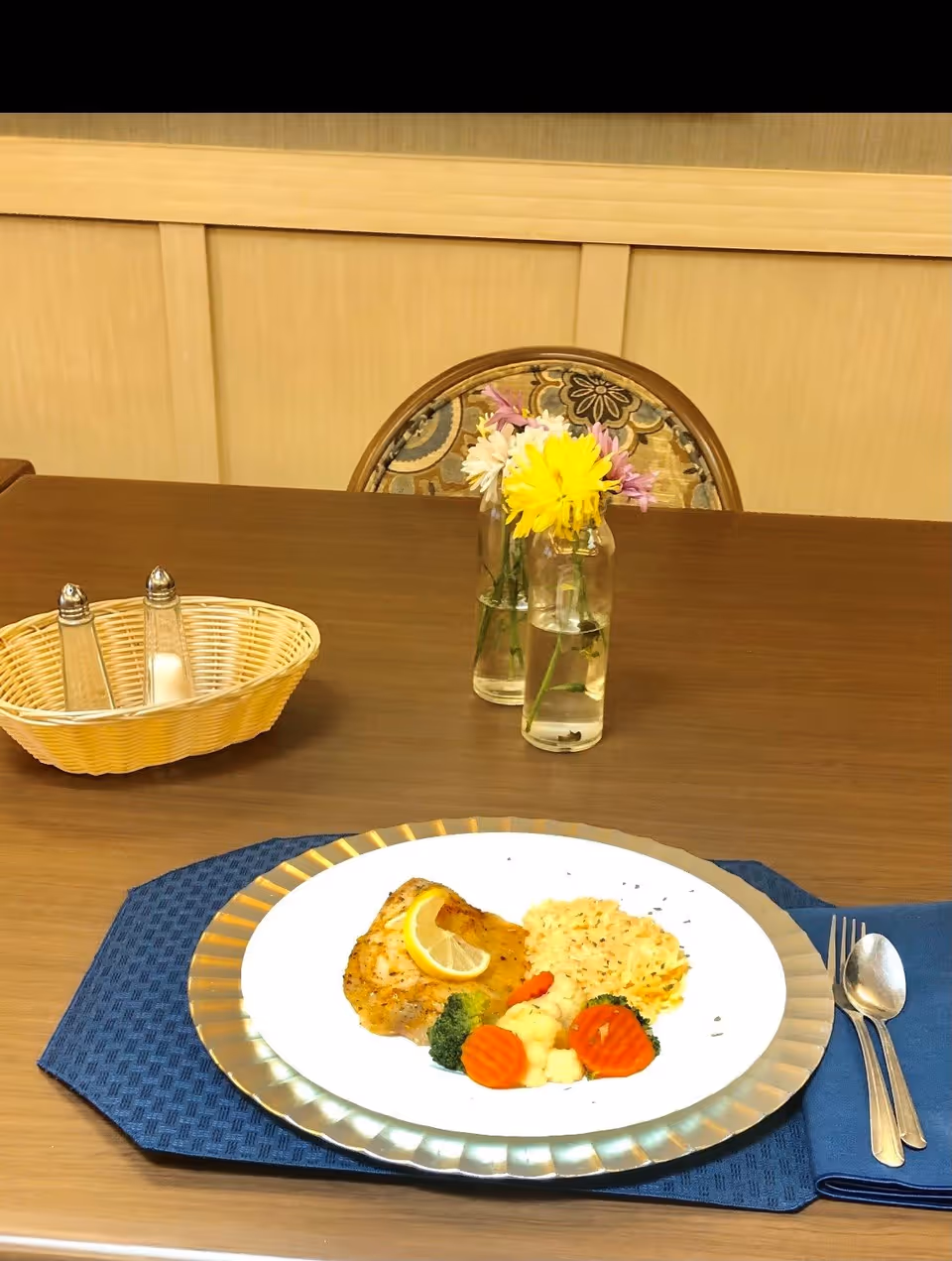 A plated meal of chicken, rice and vegetables on a place setting on a dining table with a small vase of flowers and a bread basket.
