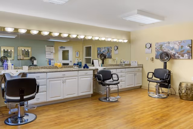 Interior view of a salon area in a senior living facility with three black salon chairs, large mirrors above white cabinets with granite countertops, hair dryers mounted on the wall, and a wooden floor. The walls are painted light yellow and decorated with floral artwork.