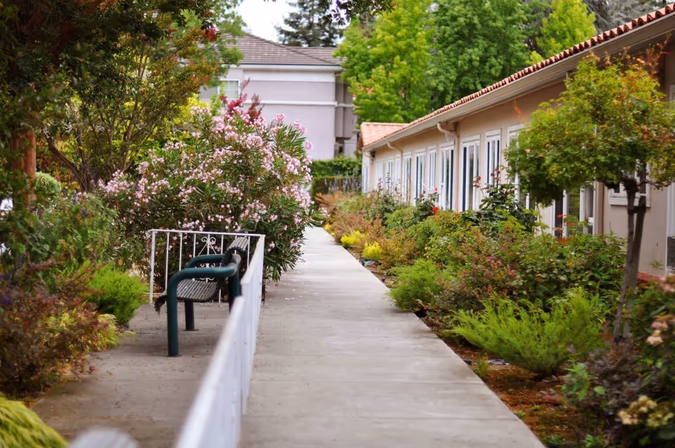 Concrete walkway between single-story residential buildings lined with flowering shrubs, trees, and a bench.