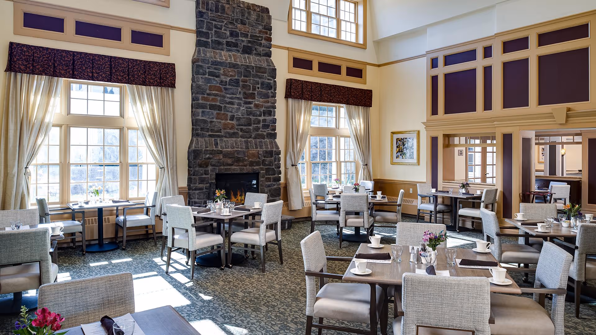 A bright and elegant dining room with multiple tables and chairs arranged neatly. The room features large windows with curtains allowing natural light to fill the space, a tall stone fireplace in the center, and decorative wall panels. Tables are set with white cups, glasses, and small flower arrangements.