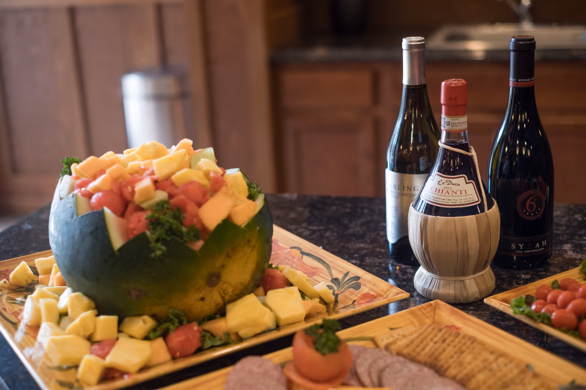 A kitchen counter with a carved watermelon bowl filled with mixed fruit pieces, surrounded by additional fruit chunks on a decorative plate. Three bottles of wine are placed behind the fruit bowl, along with plates of crackers, sliced meats, and cherry tomatoes.