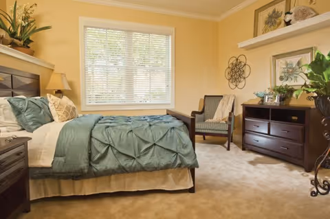 Bright, warmly painted bedroom featuring a teal-blanketed bed, armchair, dresser, and a window with blinds.
