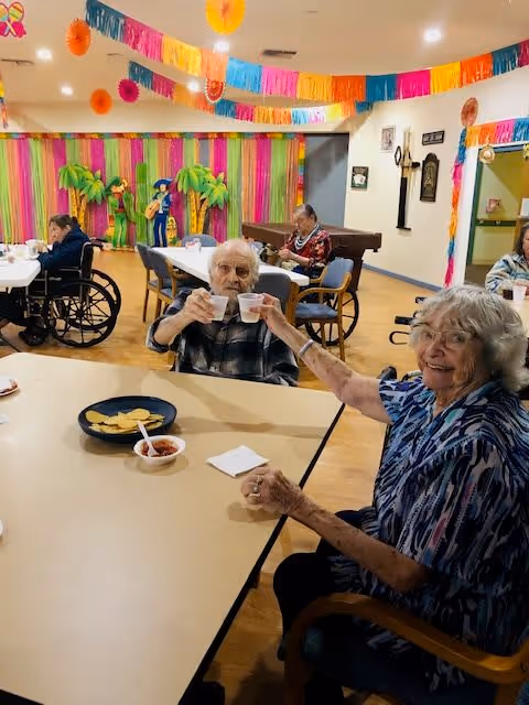 A group of elderly people sitting at tables in a decorated room with colorful streamers and paper decorations hanging from the ceiling. Two elderly individuals at the front are smiling and clinking plastic cups together. There are snacks on the table, and other seniors are seated in the background, some in wheelchairs.