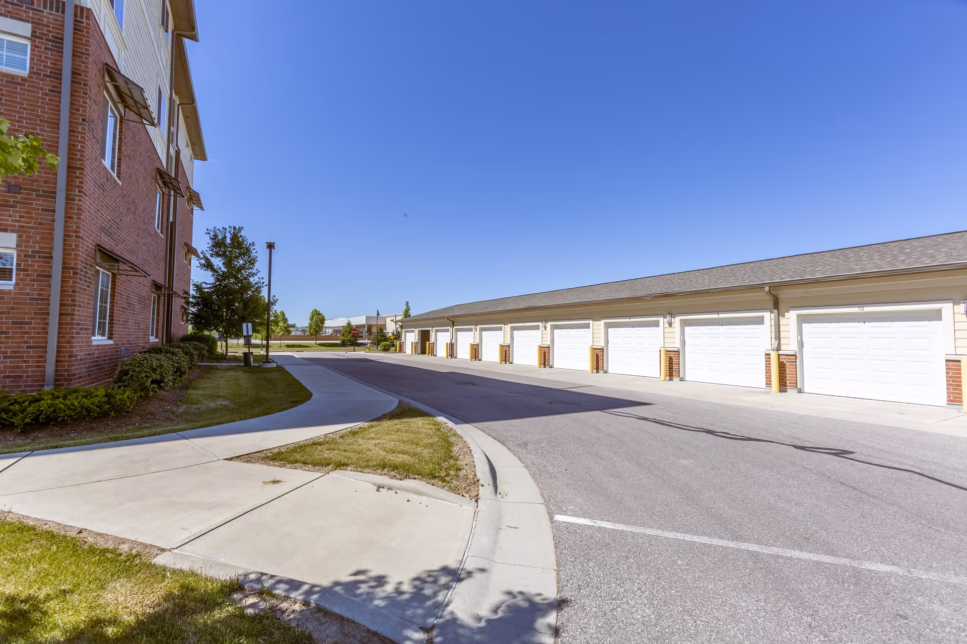 Outdoor view of a senior living facility showing a row of white garage doors on the right and a red brick building on the left under a clear blue sky. There is a paved road and sidewalk with some greenery and trees along the side.