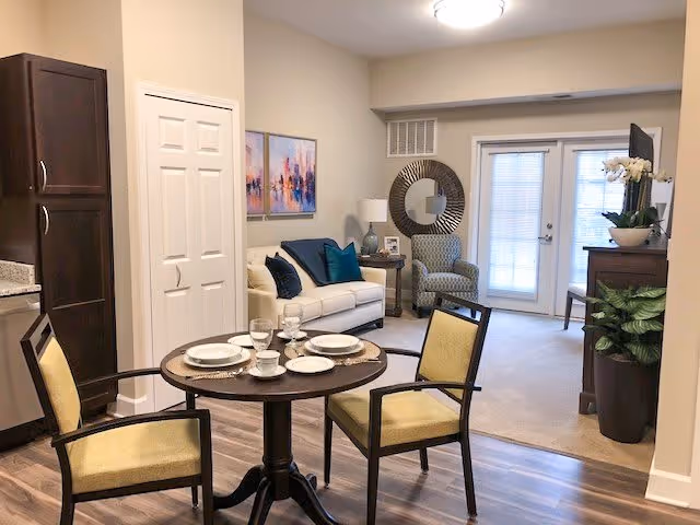 A cozy living and dining area in a senior living facility with a small round dining table set for four, a white sofa with blue and teal cushions, a patterned armchair, a round decorative mirror on the wall, and a cabinet with a potted plant and TV. The room has light-colored walls, wood flooring in the dining area, and carpet in the living area, with glass doors letting in natural light.