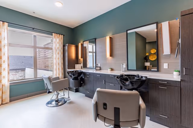 A modern hair salon area inside a facility with two salon chairs facing black wash basins and large mirrors mounted on a wall with wooden cabinetry below. The room has a large window with patterned curtains allowing natural light to enter, and the walls are painted a muted green color.