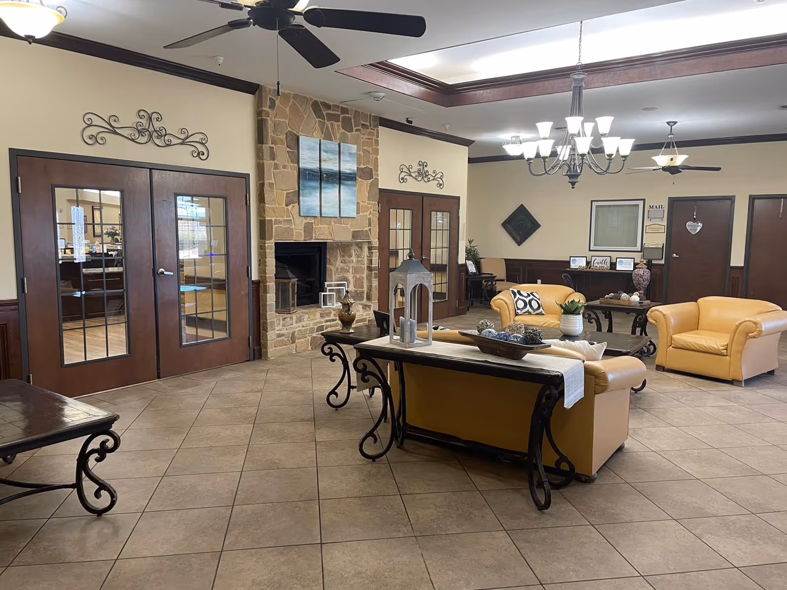 A spacious living room area in a senior living facility featuring tan leather sofas and armchairs arranged around a coffee table. The room has a stone fireplace with a three-panel abstract painting above it, large ceiling fans, a chandelier, and decorative wall accents. There are double glass doors on one side and a desk area with framed pictures and signs in the background.