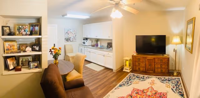 Cozy living area with a sofa, rug, TV on a wooden cabinet, dining table, and a small kitchenette in the background.
