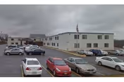 Exterior view of a two-story beige building with multiple windows, identified as Massena Rehabilitation and Nursing Center, with an American flag on a flagpole in front. Several cars are parked in the parking lot in front of the building under a cloudy sky.