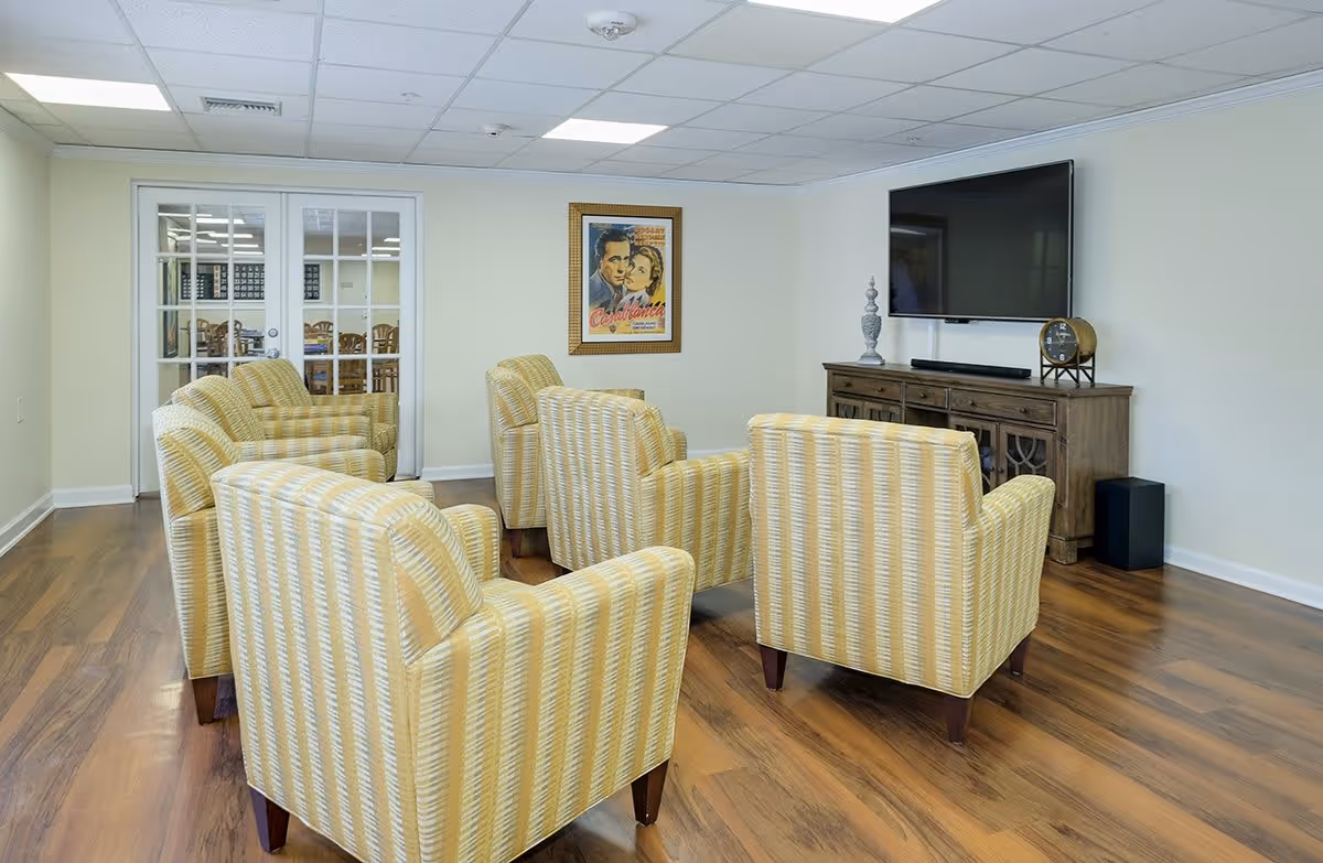 A cozy living room area with six yellow patterned armchairs arranged in two rows facing a large flat-screen TV mounted on the wall above a wooden cabinet. The room has wood flooring, a white ceiling with recessed lighting, and a framed Casablanca movie poster on the wall. Glass double doors lead to another room with tables and chairs.