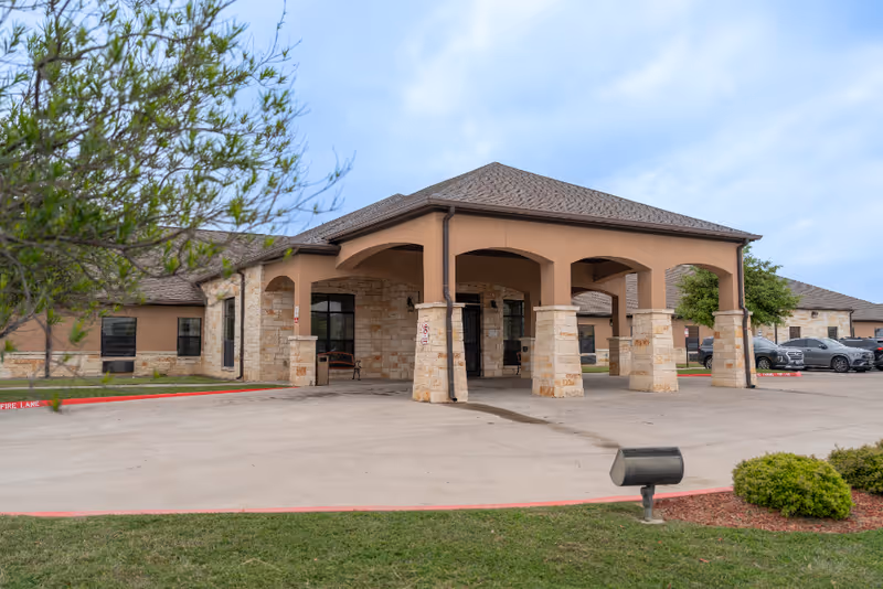 Exterior view of a single-story nursing and rehabilitation center building with a covered entrance supported by stone pillars. The building has a combination of stone and stucco walls with a shingled roof. There are parked cars visible on the right side and some greenery including bushes and a tree in the foreground.