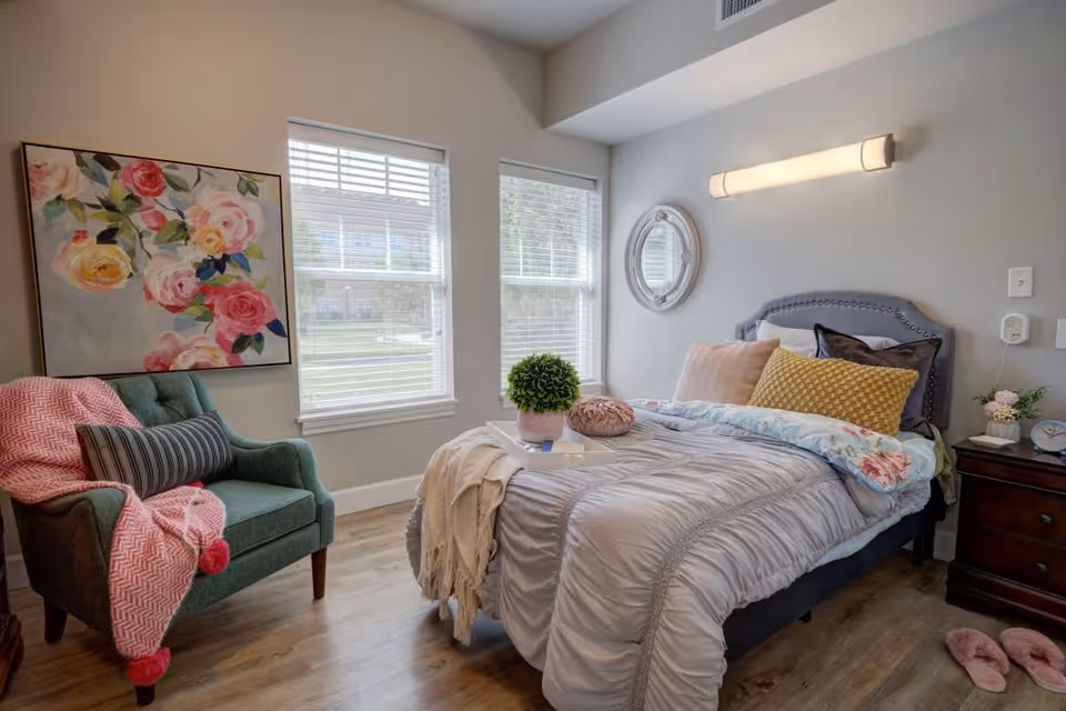 A cozy bedroom with a neatly made bed featuring multiple pillows and a floral comforter. Next to the bed is a wooden nightstand with a small plant, clock, and book. A green armchair with a pink throw blanket and striped pillow sits near two large windows with white blinds. A colorful floral painting hangs on the wall above the chair, and a round mirror is mounted on the wall above the bed. The room has wooden flooring and soft lighting.