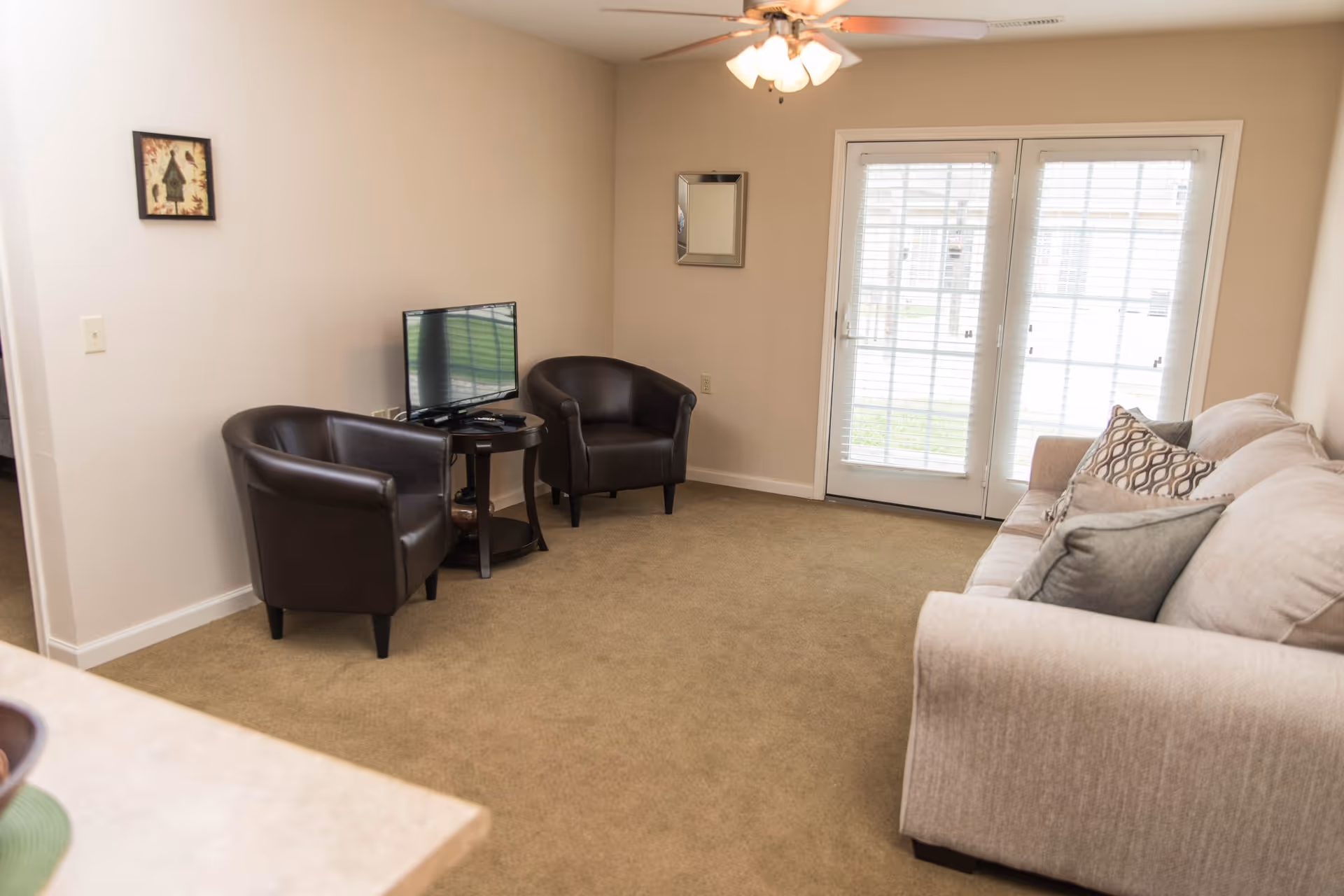A living room with beige carpet, a beige sofa with patterned and solid pillows, two dark brown leather armchairs, a small round table with a flat-screen TV on it, a ceiling fan with lights, and double glass doors with white blinds leading outside.
