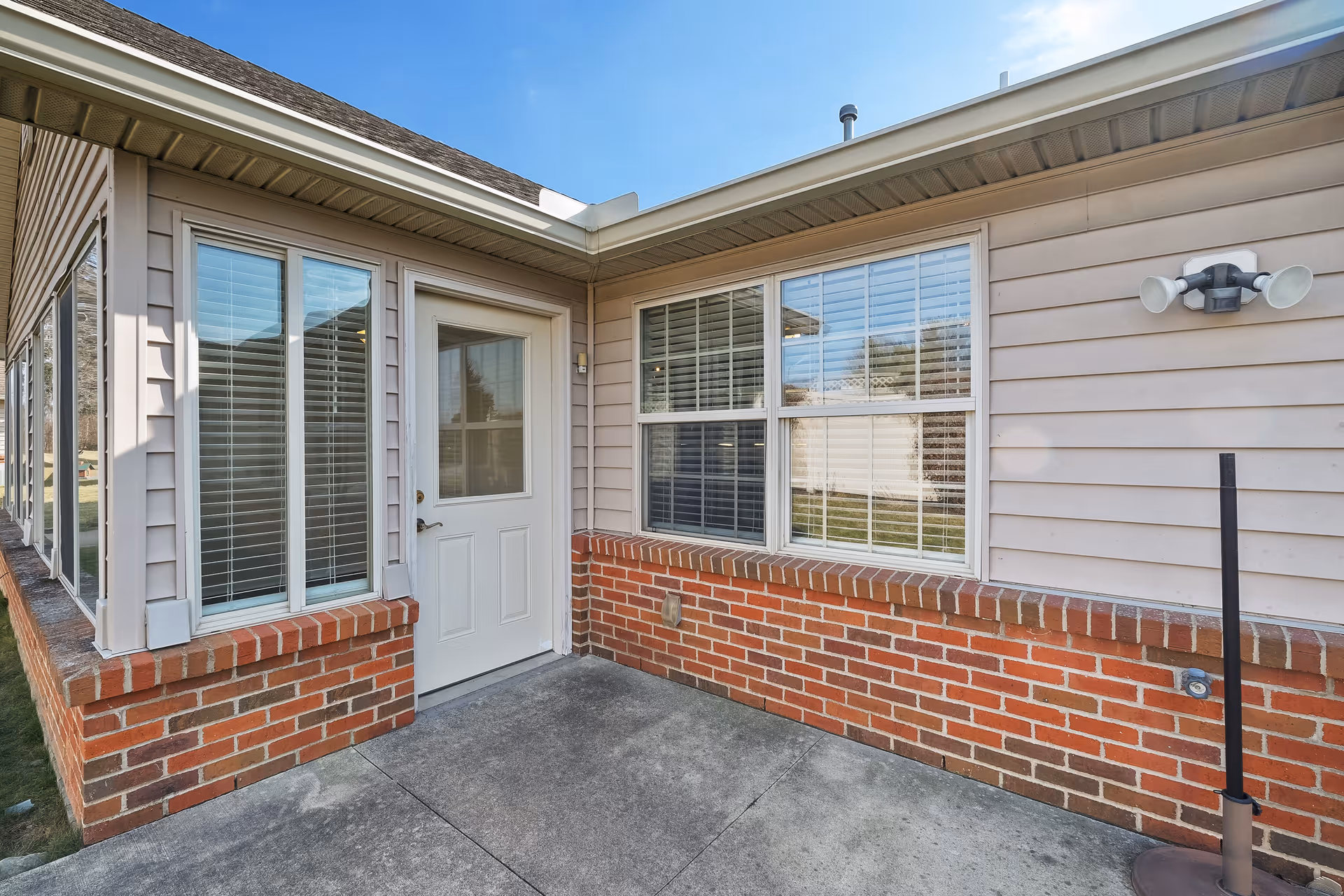 Exterior corner of a building with beige siding and red brick trim, featuring a white door with a window and two large windows with white blinds. There is a concrete patio area in front and a dual spotlight mounted on the wall.