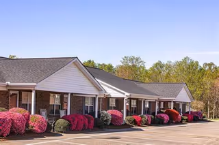 Single-story brick retirement cottages with covered porches, colorful flowering shrubs and an empty parking lot under a clear blue sky.