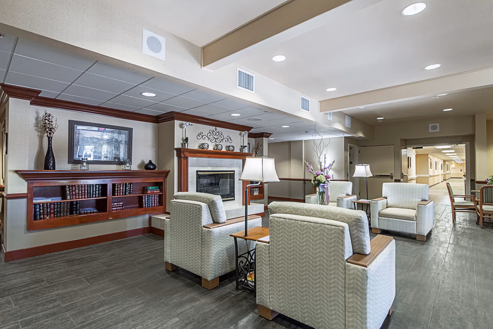 Comfortable lounge area with armchairs, side tables, lamps, a fireplace and bookshelf in a senior living facility.