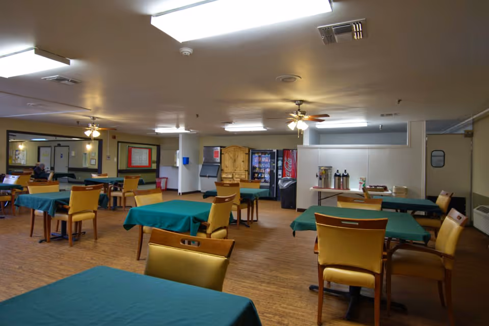 A dining room with multiple tables covered with green tablecloths and surrounded by yellow chairs. The room has a wooden floor, ceiling fans with lights, vending machines, and a table with coffee dispensers and cups against the wall.