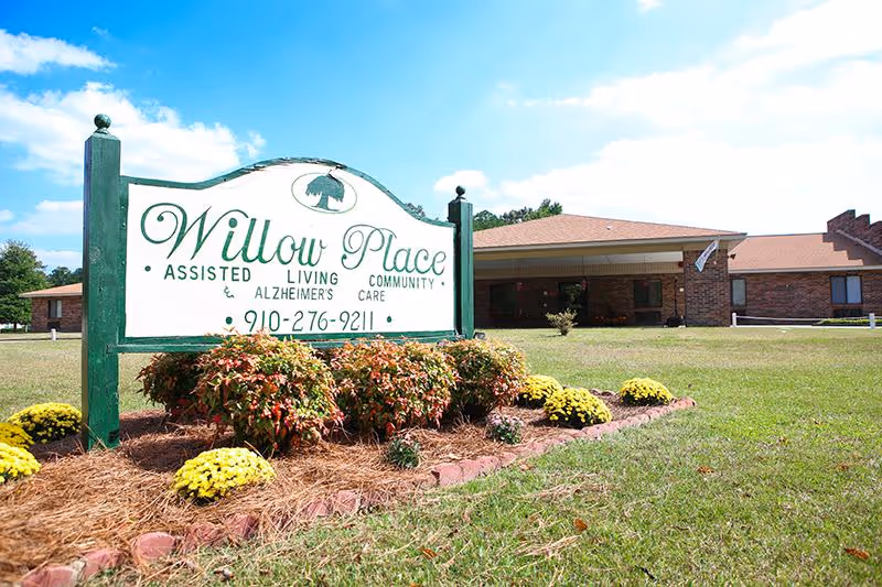 Outdoor view of Willow Place Assisted Living & Alzheimer's Care Community sign with a brick building in the background, surrounded by grass, flowers, and a clear blue sky.