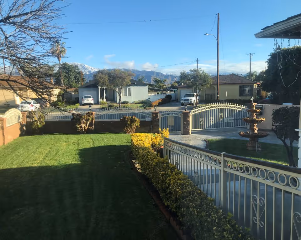View of a residential front yard with a well-maintained green lawn, yellow flowering bushes along a white metal fence, a multi-tiered fountain on the right side, and a gated driveway. In the background, there are houses, parked cars, trees, and mountains under a clear blue sky.