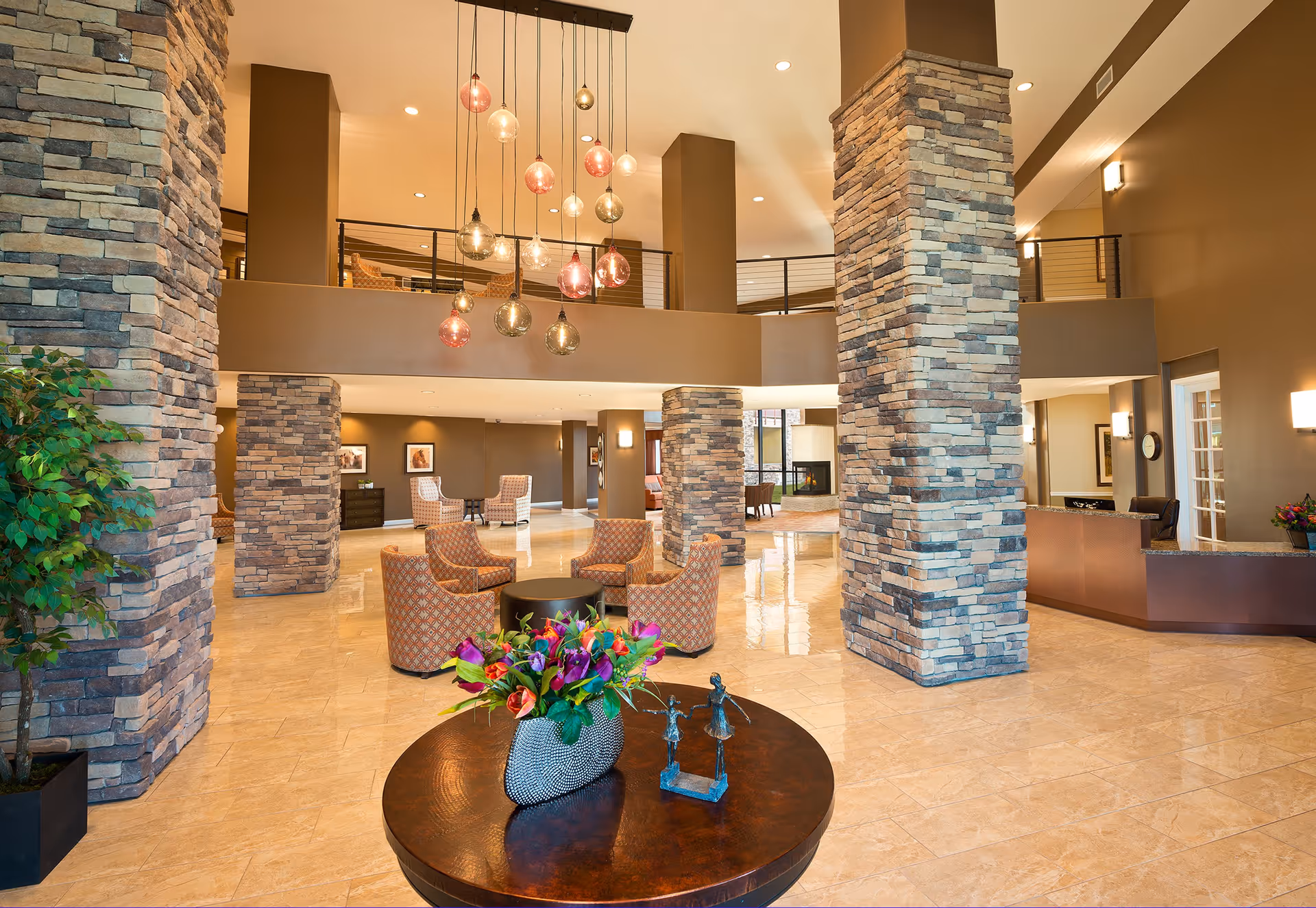 Spacious and well-lit lobby area with stone pillars, a round wooden table with a vase of colorful flowers and a small sculpture, several patterned armchairs arranged around small tables, modern hanging pendant lights, and a reception desk on the right side.