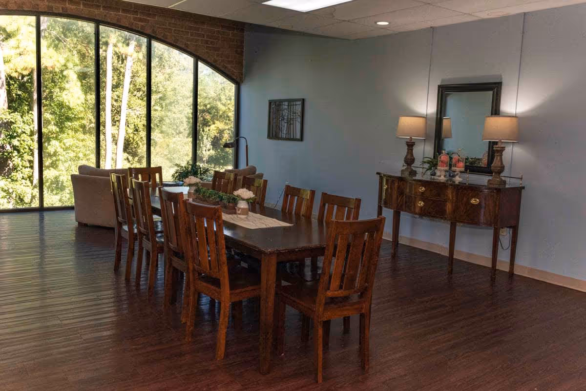 A spacious dining room with a long wooden table surrounded by eight wooden chairs. The room has large floor-to-ceiling windows with an arched top, allowing natural light to illuminate the space and providing a view of green trees outside. Against one wall, there is a wooden sideboard with two lamps, a mirror, and decorative items. The floor is made of dark wood, and the walls are painted light blue.