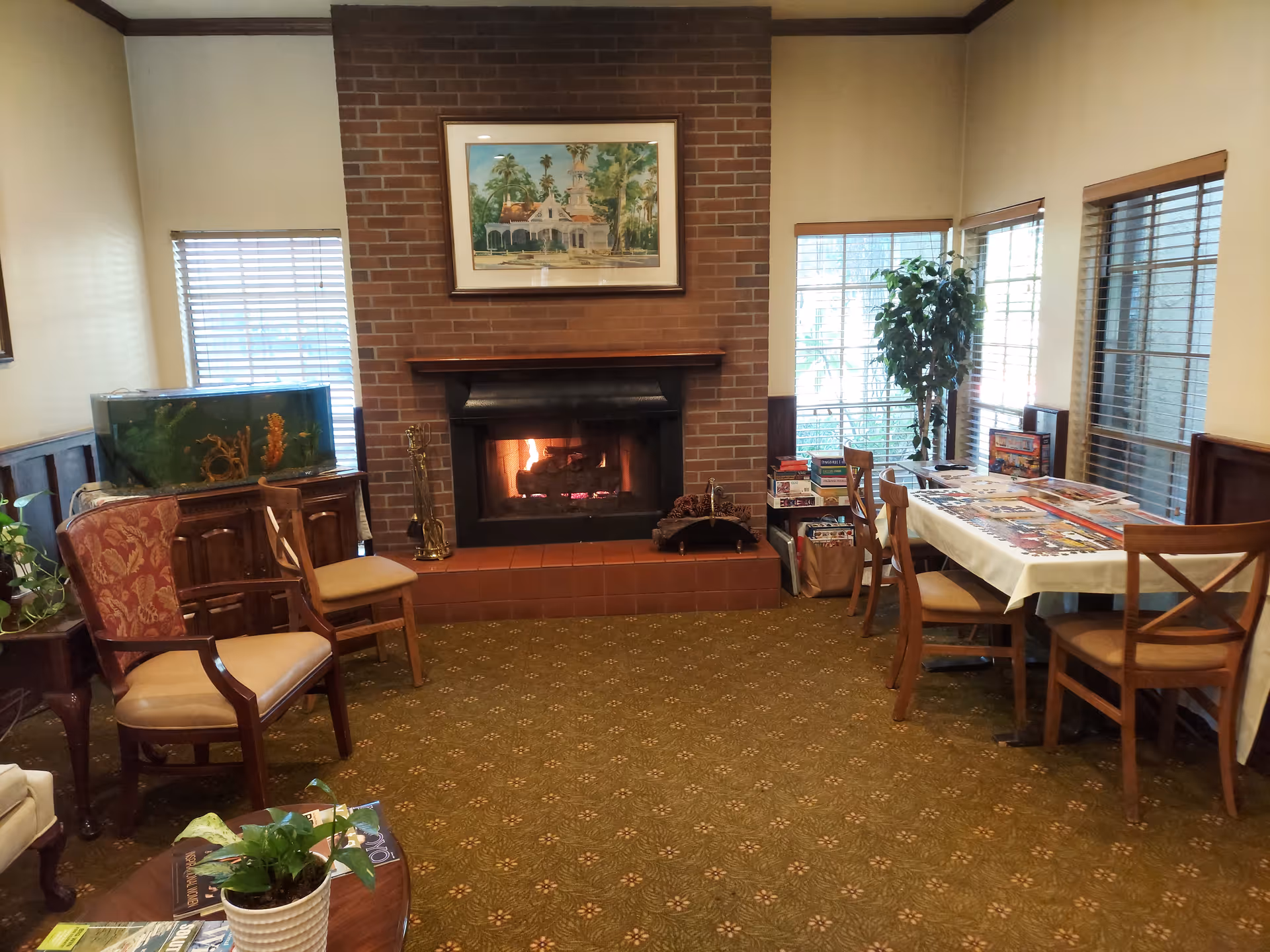 Cozy common room with a lit brick fireplace centered on the back wall. Above the fireplace hangs a framed painting of a house with palm trees. To the left of the fireplace is a fish tank on a wooden cabinet and two upholstered chairs. To the right is a table covered with a white tablecloth, surrounded by wooden chairs, with board games and puzzles on top and stacked nearby. Large windows with blinds let in natural light, and there are several potted plants around the room. The floor is carpeted with a patterned design.