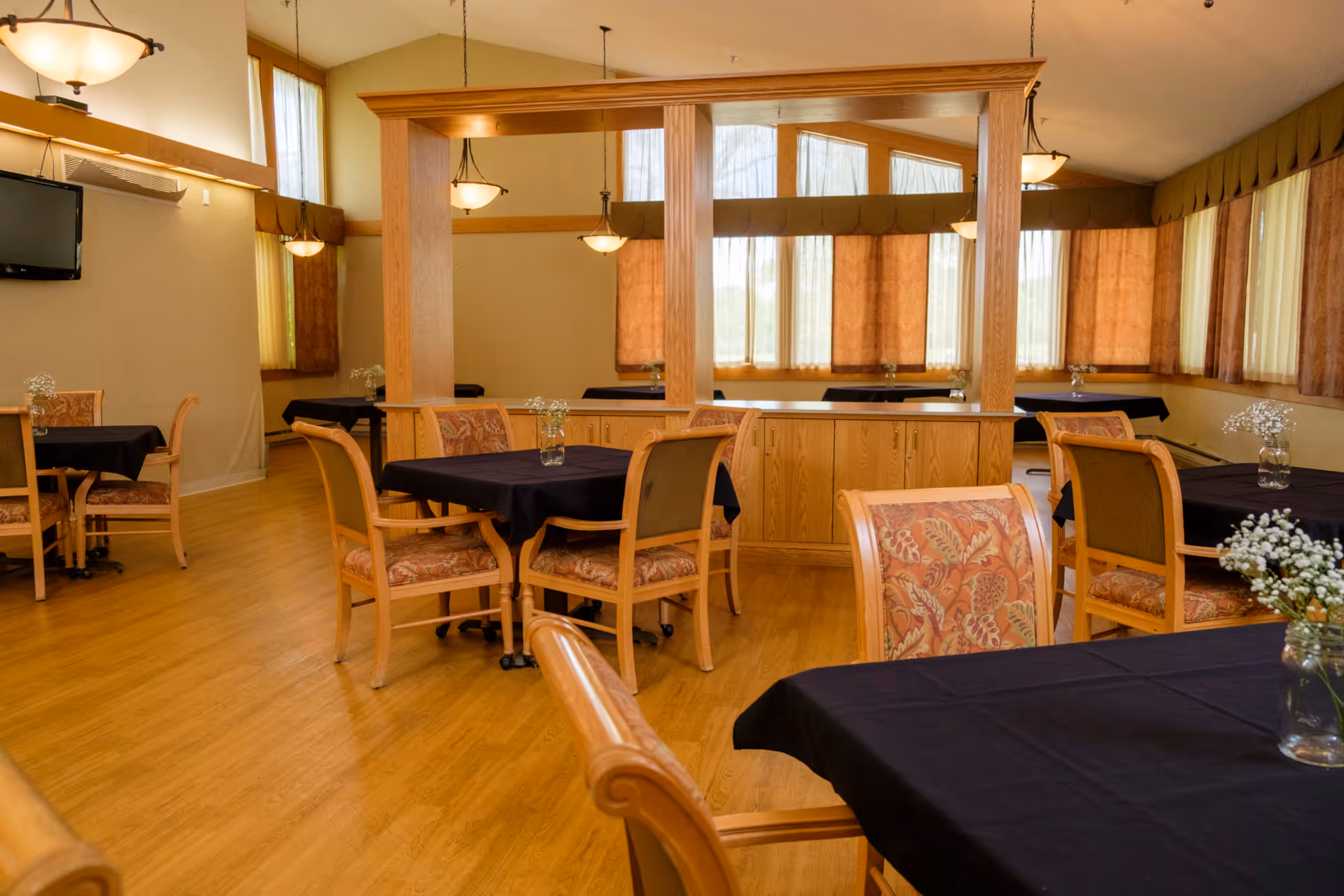 A dining room with several wooden tables covered with black tablecloths and surrounded by cushioned chairs with floral upholstery. The room has large windows with sheer and brown curtains, wooden flooring, and hanging light fixtures. There is a wooden partition with cabinets in the center of the room and a wall-mounted TV on the left side.
