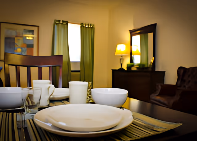 A dining table set with white plates, bowls, glasses, and cups in a room with green curtains, a wooden chair, a dresser with a mirror, and a lit table lamp.