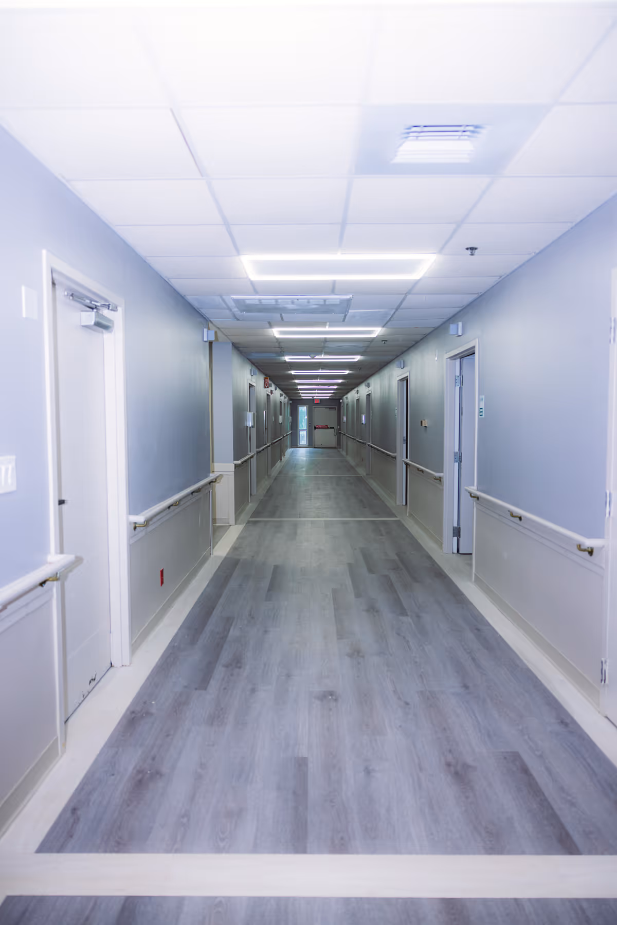 A long, well-lit hallway in a healthcare or rehabilitation facility with light gray walls, handrails on both sides, multiple closed doors, and a light gray wood-patterned floor.