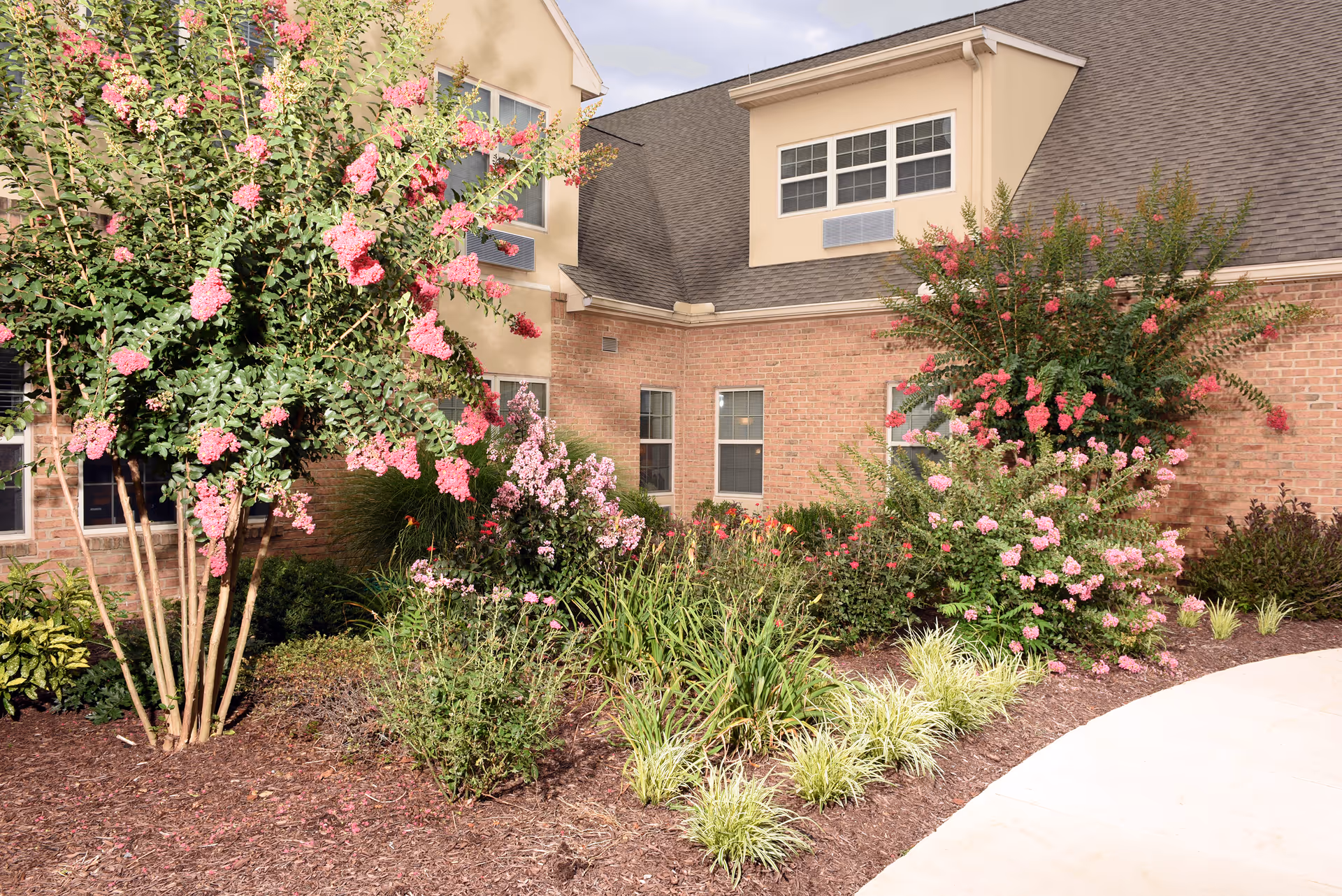 Exterior view of a brick building with beige siding and multiple windows, surrounded by landscaped garden beds featuring pink flowering bushes, green shrubs, and a curved concrete walkway.