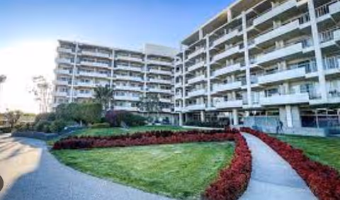 Exterior view of a multi-story senior living facility with balconies, surrounded by well-maintained green lawns and landscaped flower beds with red flowers along a curved walkway.