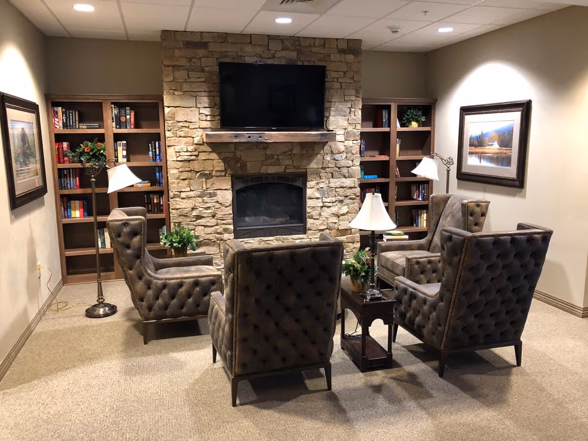 A cozy living room area with four tufted armchairs arranged around a small wooden table with a lamp and plants. The room features a stone fireplace with a mounted flat-screen TV above it, two wooden bookshelves filled with books and decorative items, and framed landscape paintings on the walls.