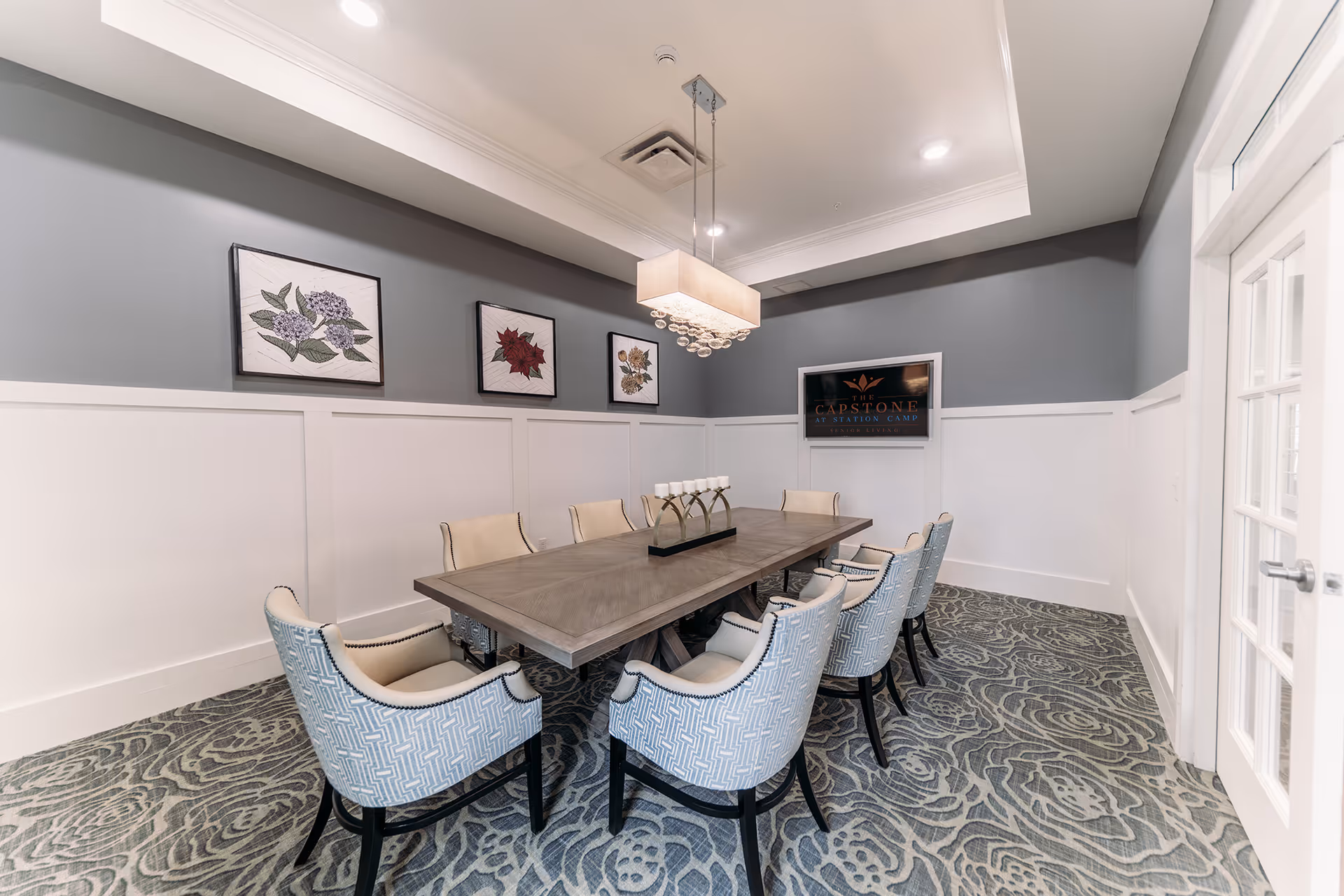 A modern dining room with a long rectangular wooden table surrounded by ten upholstered chairs with patterned fabric. The room has gray walls with white wainscoting, three framed floral artworks on one wall, a decorative chandelier hanging above the table, and a TV mounted on the wall displaying The Capstone at Station Camp logo. The floor is covered with a patterned carpet, and there is a glass door on the right side.