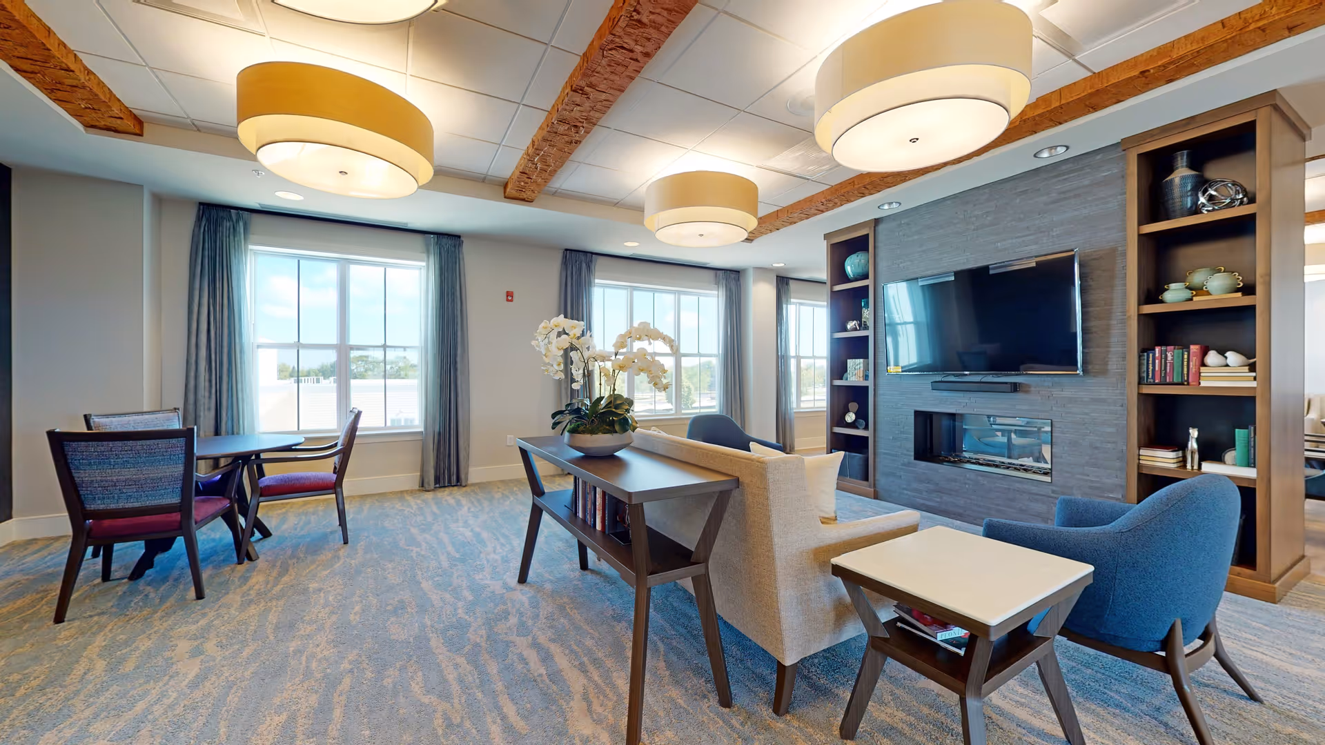 Bright assisted living common lounge with seating, a TV over a fireplace, bookshelves, tables, and large windows.