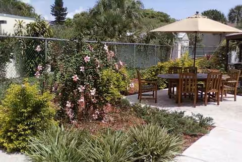 Outdoor patio with wooden table and chairs, umbrella, surrounded by greenery and flowers.