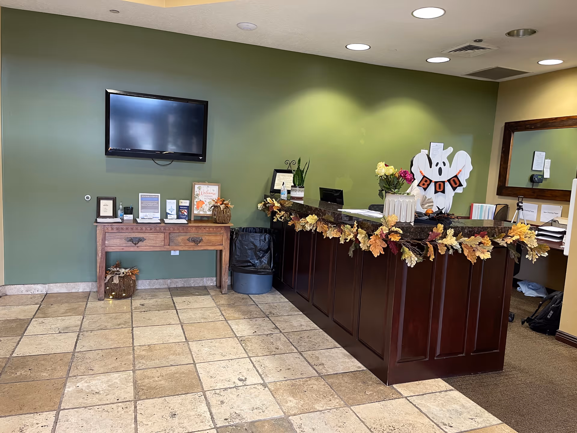 Reception area with a dark wooden front desk decorated with autumn leaves garland and a ghost cutout with a 'BOO' banner. Behind the desk are flowers, office supplies, and a mirror on the wall. To the left, a green wall with a mounted flat screen TV above a wooden table holding framed certificates and decorations. The floor is tiled with beige stone tiles.