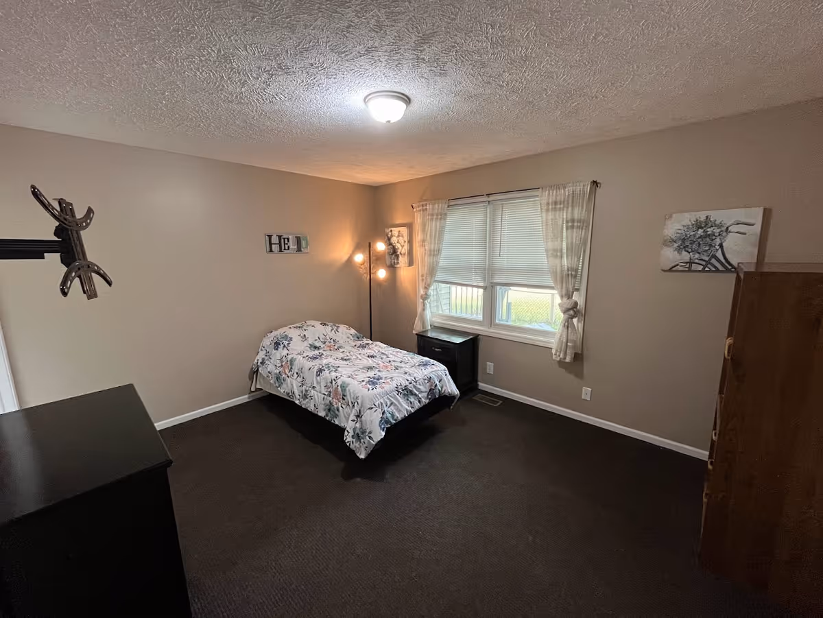 A simple bedroom with beige walls and dark carpet. The room contains a single bed with floral bedding, a black nightstand under a window with white curtains, a floor lamp with multiple bulbs, a wooden dresser, and a wall-mounted TV bracket. There are two pieces of wall art, one above the bed and one near the dresser.