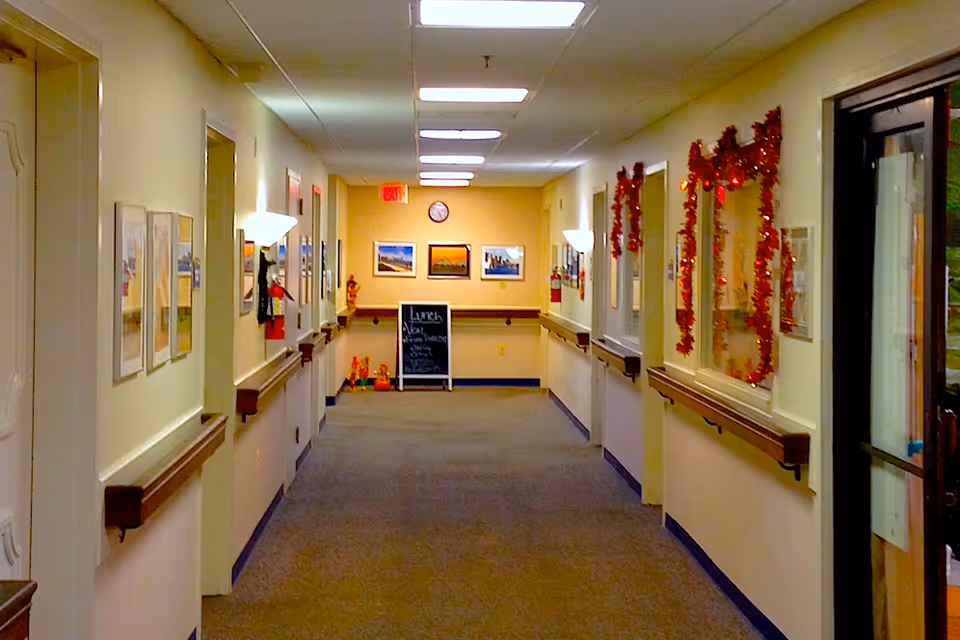 Well-lit decorated hallway in an assisted living facility with handrails, framed pictures, garlands, and a small chalkboard at the far end.