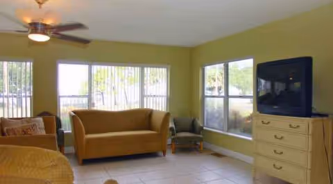 Bright living room with yellow walls, seating, large windows, and a TV on a dresser.