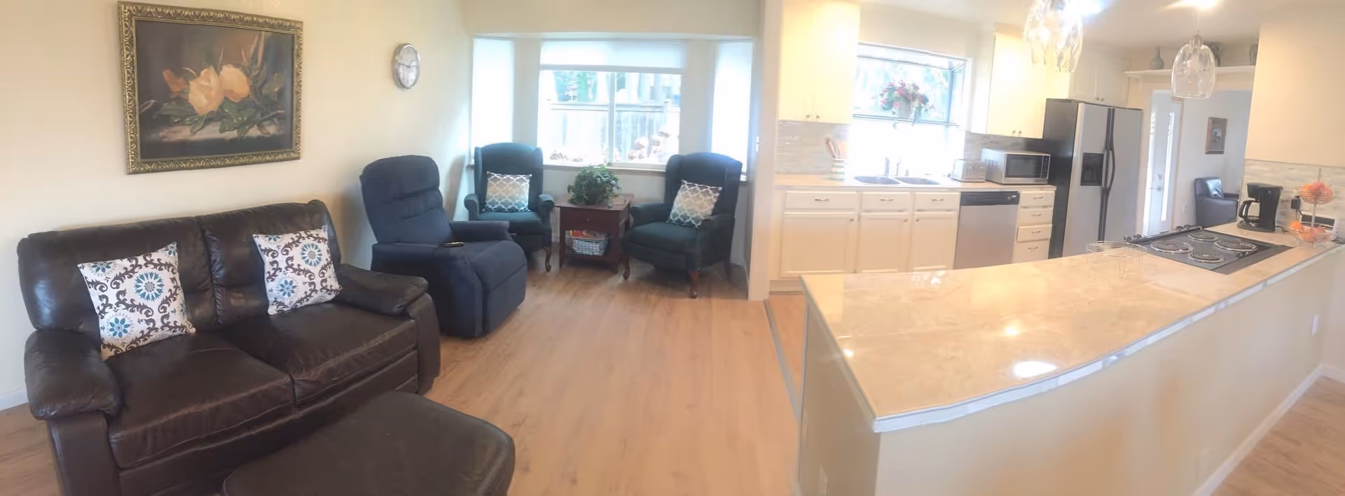 A bright and cozy living room and kitchen area in a senior living facility. The living room features a dark brown leather sofa with two patterned pillows, two blue armchairs with pillows, a recliner, a small wooden side table with a plant, and a large window letting in natural light. The kitchen has white cabinets, a double sink, a microwave, a stainless steel refrigerator, a coffee maker, and a countertop with an electric stove and two pendant lights hanging above.