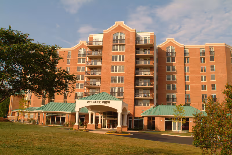 Front exterior view of a multi-story brick building with balconies and a green roof entrance canopy labeled '211 PARK VIEW', surrounded by grass, trees, and a clear sky.