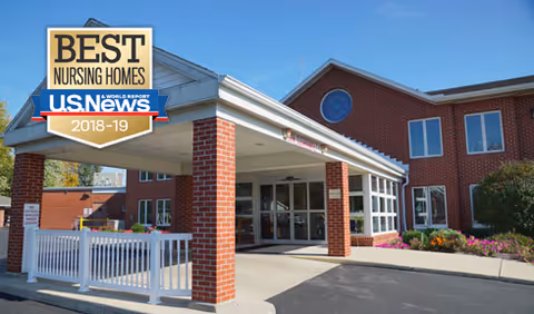 Entrance of a brick building with a covered drop-off area supported by brick columns and white railings. There are windows and landscaping with flowers near the entrance. A badge in the top left corner reads 'Best Nursing Homes U.S. News 2018-19'.