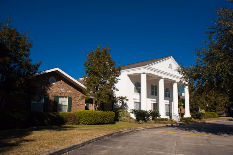 Front exterior of a white, columned building with a brick wing, driveway, and landscaping under a clear blue sky.