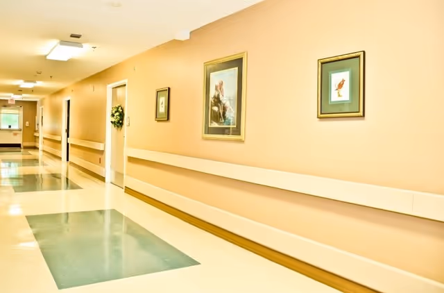 Well-lit beige interior hallway with handrails, framed artwork, doors, and green tile floor accents.