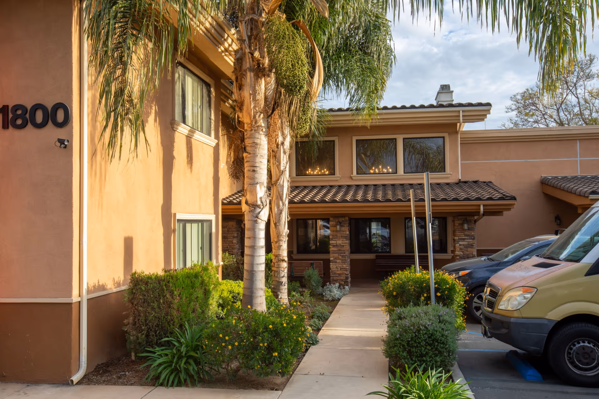 Exterior view of Mainplace Senior Living building showing a walkway lined with palm trees and bushes leading to the entrance. Several parked vehicles are visible on the right side, and the building has a tan stucco finish with stone accents and a tiled roof.