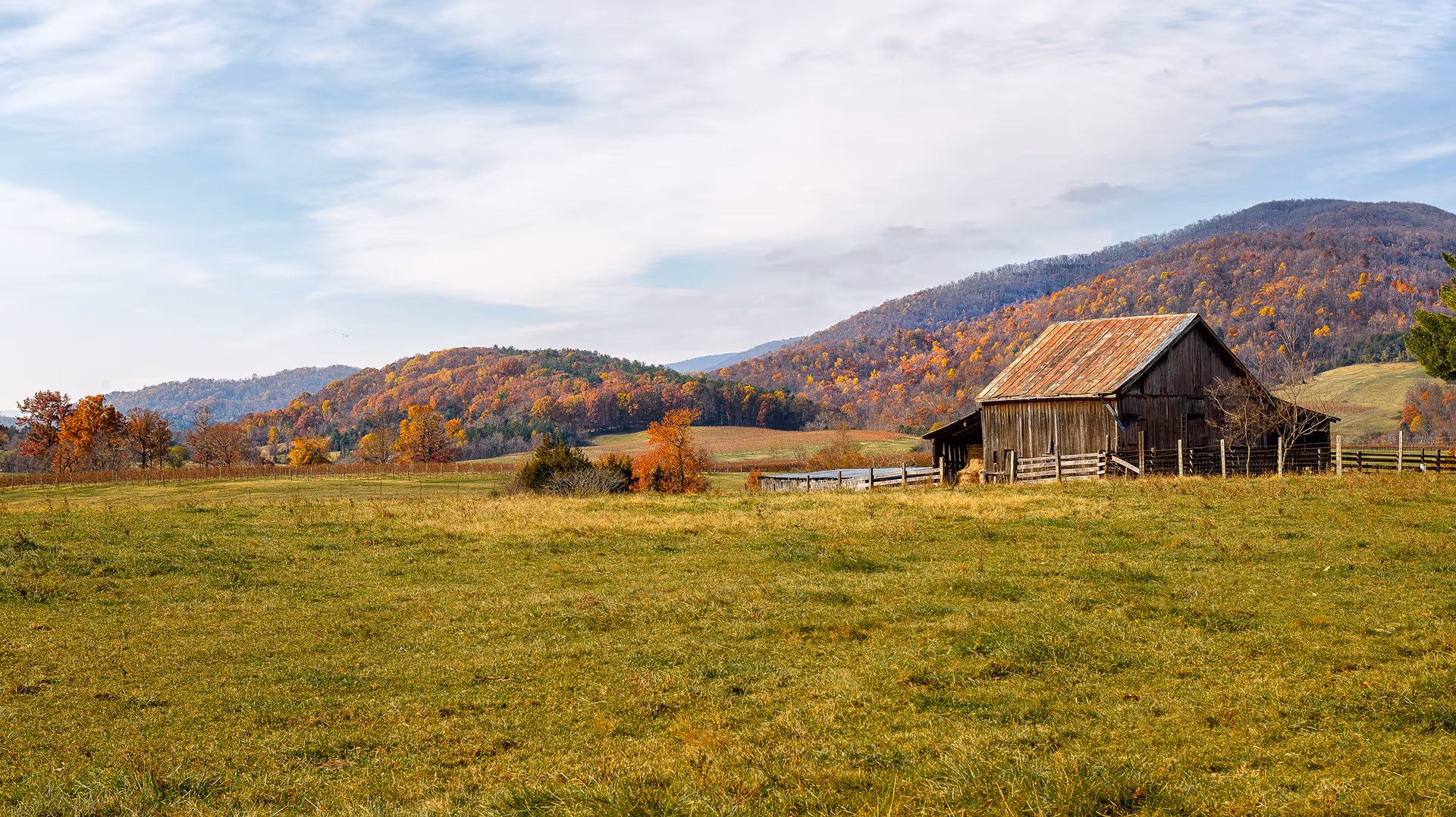 A weathered barn sits in a grassy field with rolling autumn hills behind.