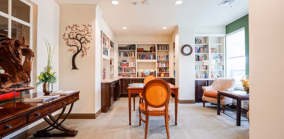 Well-lit communal reading room with bookshelves, a central table and chairs, and comfortable seating by a window.