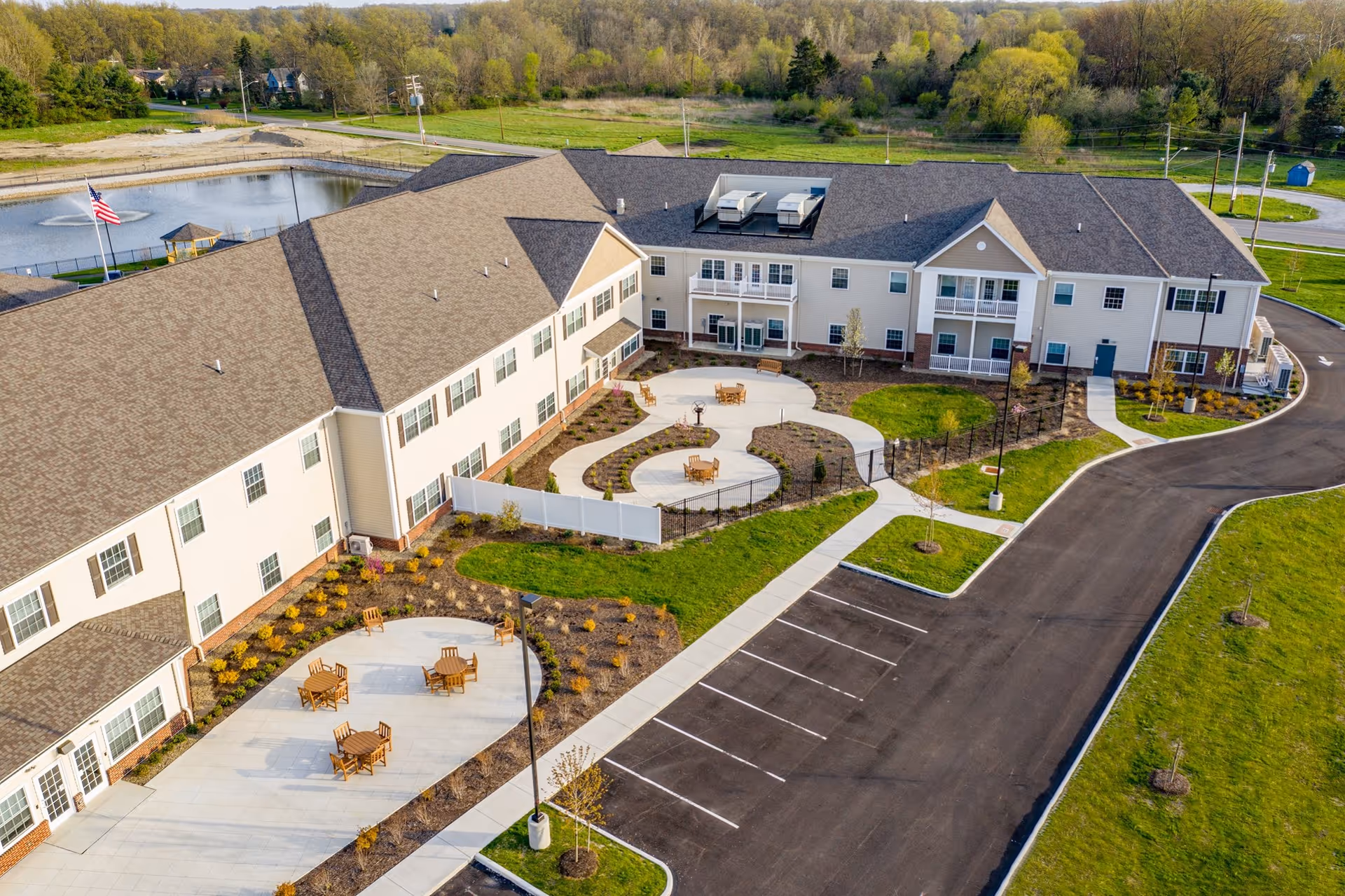 Aerial view of a two-story senior living building with landscaped courtyards, outdoor seating, parking lot, and nearby pond.